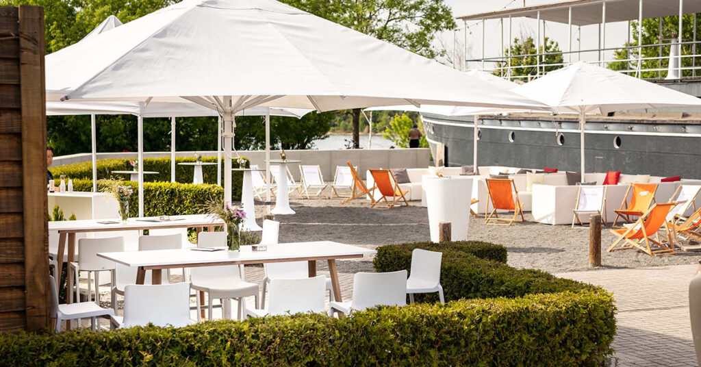Der Außenbereich der amSee Eventlocation: Weiße Loungemöbel und Liegestühle in orange sowie weiße Sonnenschirme stehen vor dem Schiffsbug in der Sonne.