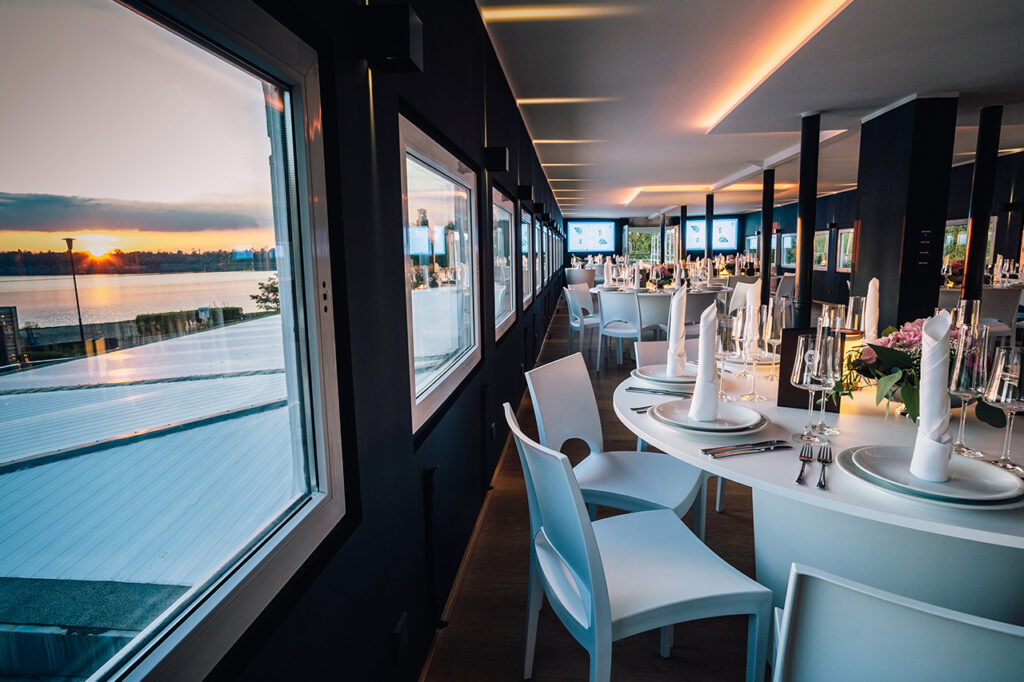 Blick an der westlichen Innenseite des Oberdecks entlang: Innen befinden sich festlich gedeckte Tische, außen leuchtet der See im Sonnenuntergang.