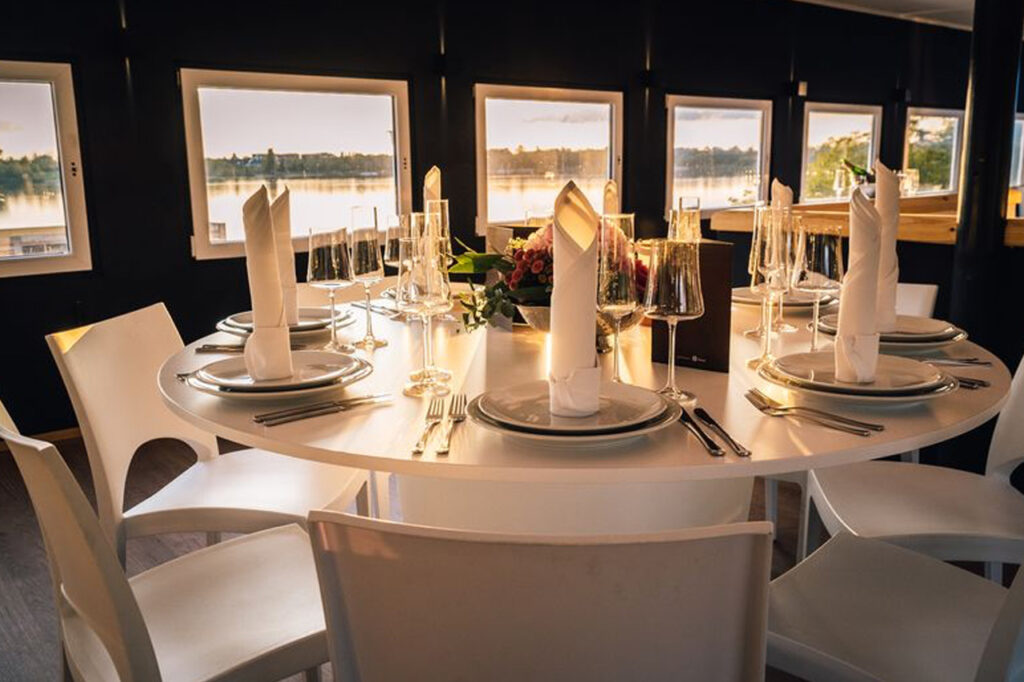 Blick ins festlich eingedeckte Oberdeck der amSee Location Leipzig, im Hintegrund ist durch die Fenster der im Sonnenuntergang glitzernde Kulkwitzer See sichtbar.