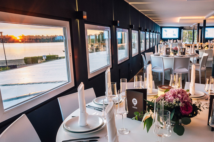 Das Bild zeigt das Oberdeck der Eventlocation amSee, dass für ein Bankett festlich gedeckt ist. Auf der linken Seite öffnet sich der Blick über den See und die untergehende Sonne.