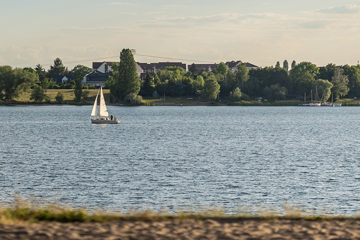 Das Bild zeigt den Blick vom Strand vor der Location amSee in Richtung See. Ein Segelboot ist darauf unterwegs.