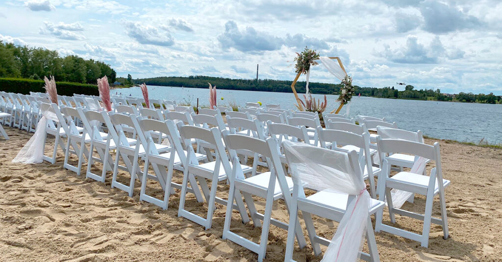 Das Bild zeigt das Setting für eine freie Trauung amSee auf der Sandterrasse: weiße Vintage-Stühle und ein hölzerner, geschmückter Traubogen.