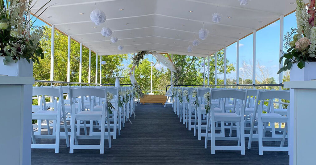 Das Bild zeigt das Setting für eine freie Trauung amSee auf dem Oberdeck: weiße Vintage-Stühle, weiße Blumensäulen und ein hölzerner, geschmückter Traubogen.