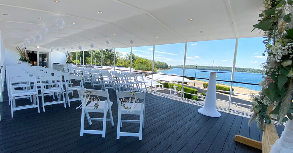 Das Bild zeigt das Setting für eine freie Trauung amSee auf dem Oberdeck: weiße Vintage-Stühle, weiße Blumensäulen und ein weißer Stehtisch für Trauredner*innen.