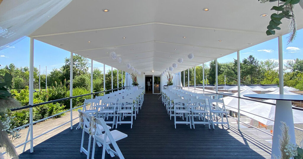 Das Bild zeigt das Setting für eine freie Trauung amSee auf dem Oberdeck: weiße Vintage-Stühle und weiße Blumensäulen.