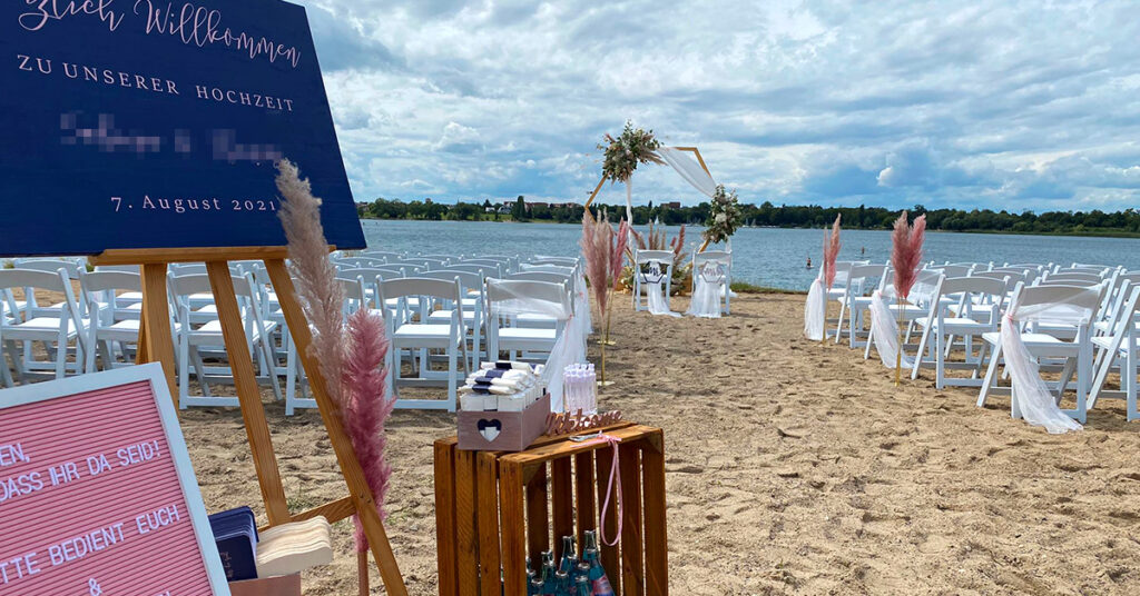 Das Bild zeigt das Setting für eine freie Trauung amSee auf der Sandterrasse: weiße Vintage-Stühle, eine hölzerne Staffelei und ein hölzerner, geschmückter Traubogen.