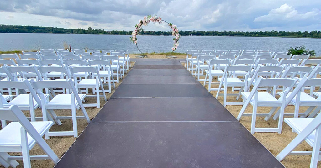 Das Bild zeigt das Setting für eine freie Trauung amSee auf der Sandterrasse: weiße Vintage-Stühle, dunkle Bodenplatten und ein hölzerner, geschmückter Traubogen.