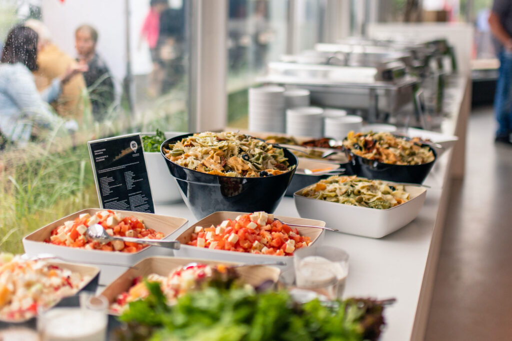 Buffet im Wintergarten, im Vordergrund Schüsseln und Platten mit Salaten, im Hintergrund Chafing Dishes.