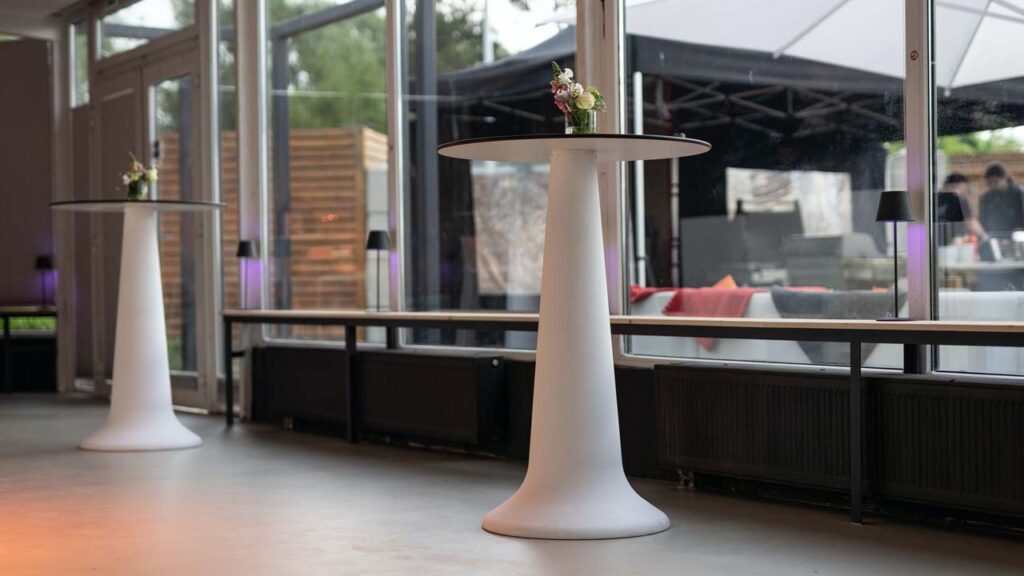 Stehtisch und Sitzbänke vor den großen Fenstern des Wintergartens, im Hintergrund der Außenbereich im Sonnenschein.