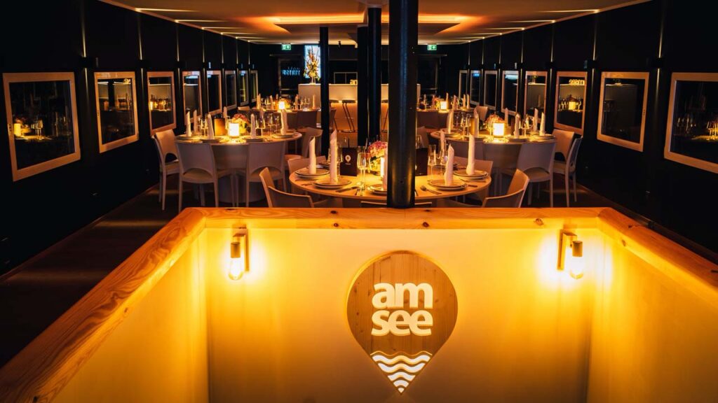 Das Oberdeck der amSee Location am Abend - im Vordergrund der beleuchtete Treppenaufgang mit amSee-Logo, im Hintergrund die Banketttische mit Ambientebeleuchtung für Winter-Events amSee.