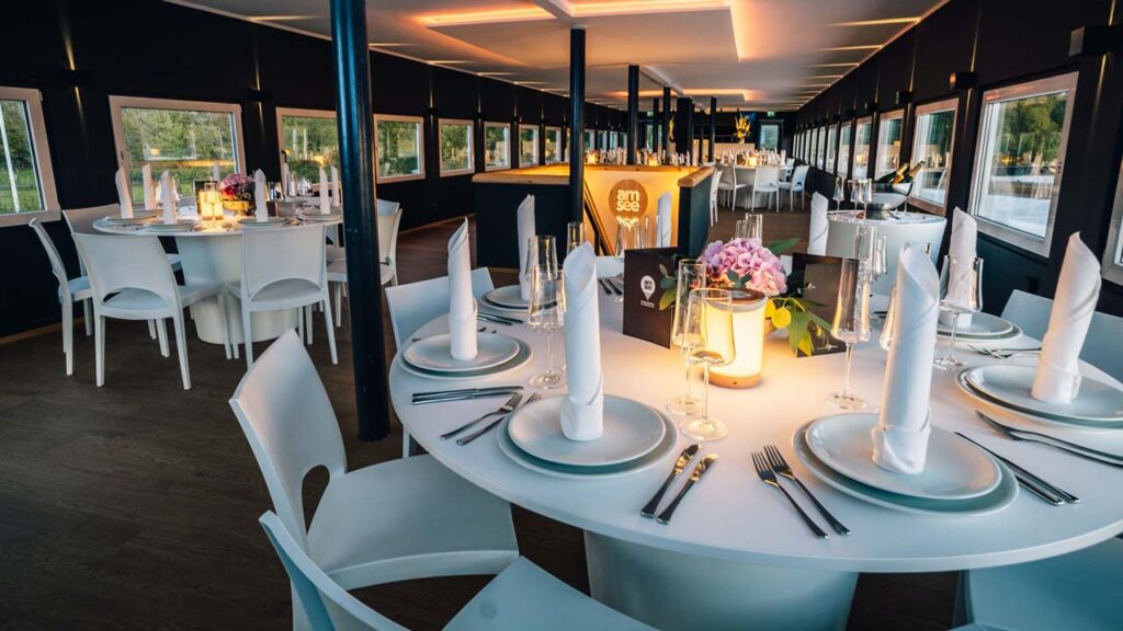 Das Oberdeck der Eventlocation amSee ist eingerichtet mit weißen modernen Bankettmöbeln. Auf den Tischen stehen Blumenschmuck, Geschirr, Gläser sowie Stoffservietten gefaltet als Kerze.