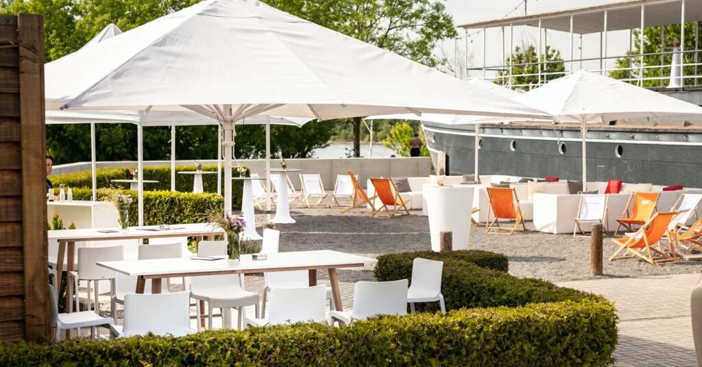 Außenbereich der Location mit weißen Sonnenschirmen, Sitzgruppen und orangefarbenen Liegestühlen. Im Hintergrund der dunkelgrau gestrichene Bug des Schiffs.