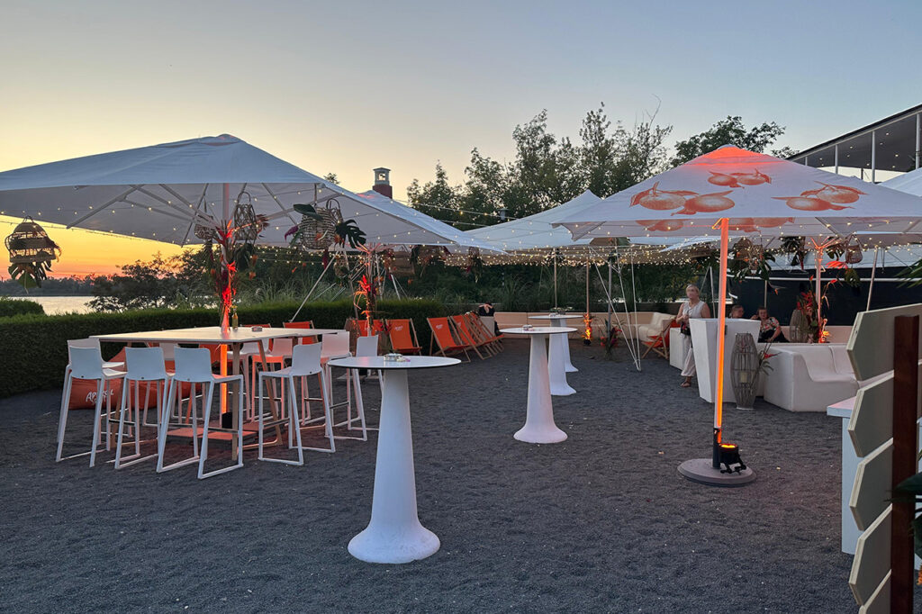 Sitzgruppen im Außenbereich vor dem Bug der amSee Location kurz nach Sonnenuntergang. In den Sonnenschirmen leuchten die ersten Lichterketten.