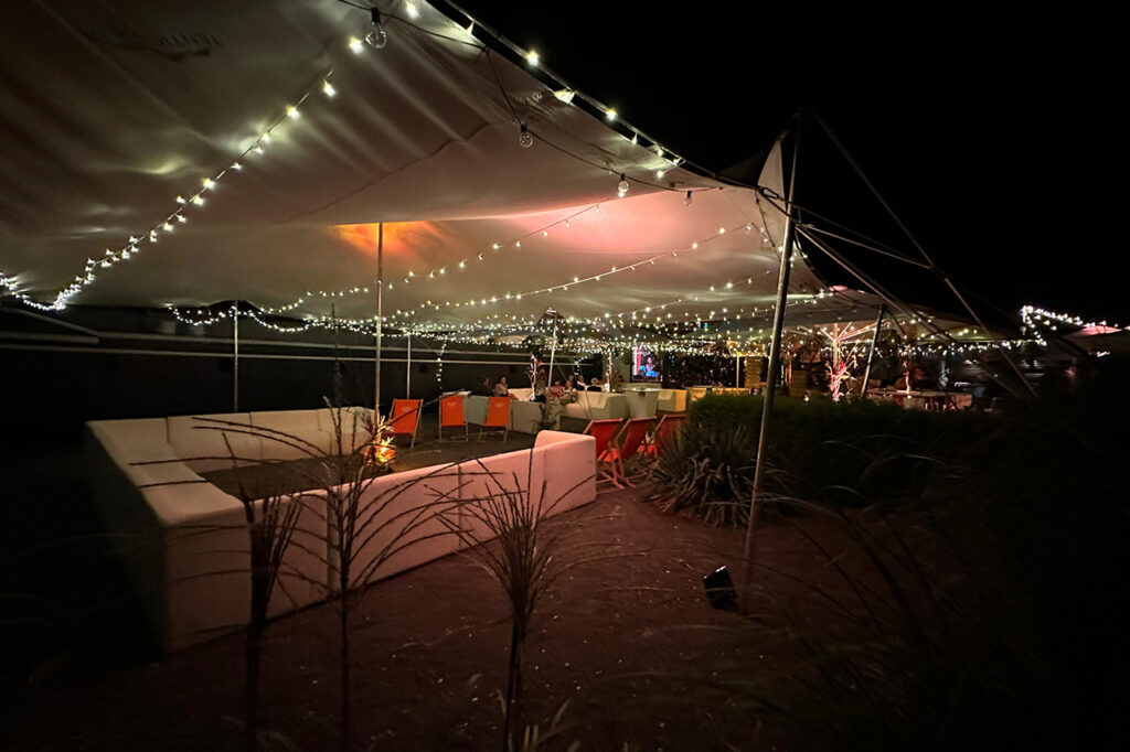 Sitzgruppen im Außenbereich der amSee Location unter einem großen Segel. Es ist Nacht, das Segel wird von mehreren Lichterketten beleuchtet.