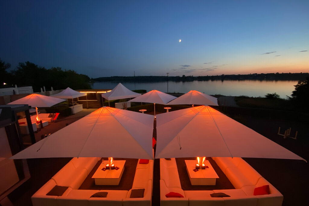 Blick über den Außenbereich der Eventlocation amSee am Kulkwitzer See während der blauen Stunde. Die weißen Möbel und Schirme werden in Amber beleuchtet.