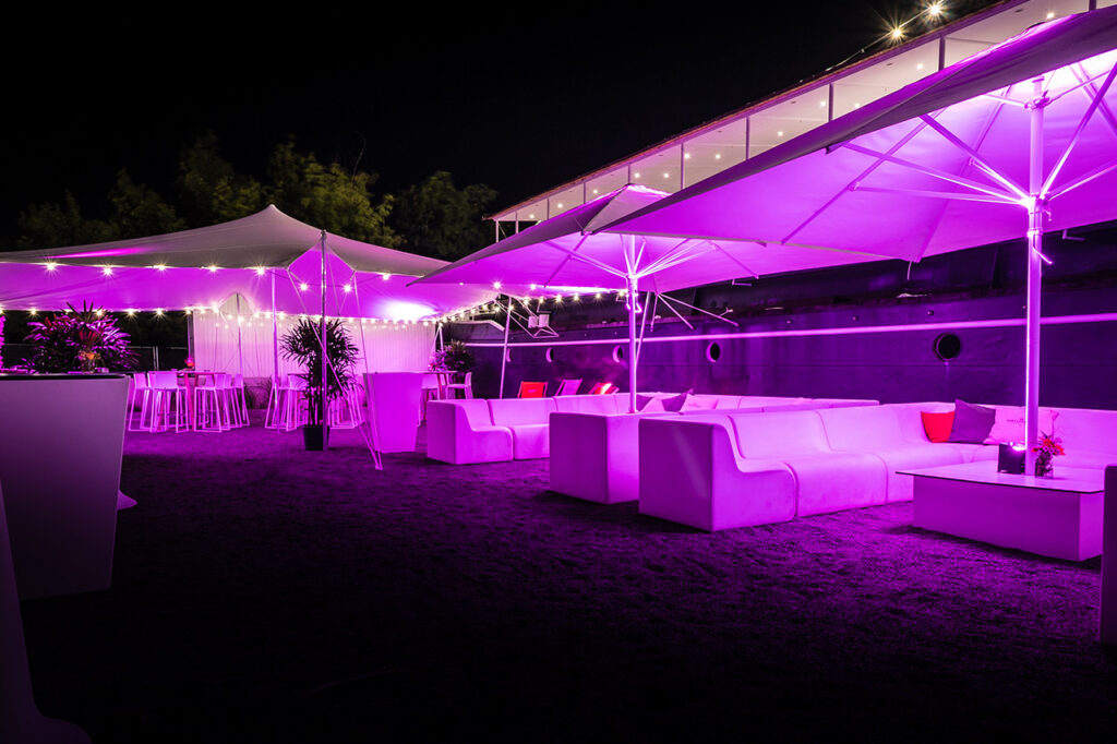 Außenbereich der Location mit Sitzgruppen unter Schirmen und Sonnensegel. Lilafarbene Beleuchtung sorgt für besondere Akzente am Abend.