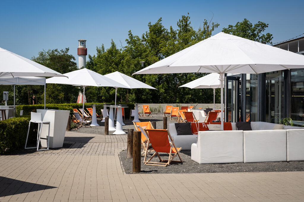 Blick auf Liegestühle und Sitzgruppen im Außenbereich der amSee Location, im Hintergrund der rot-weiße Leuchtturm vor der Location.