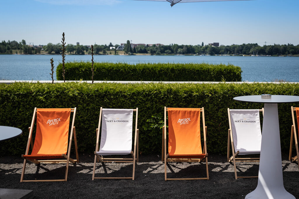 Blick auf Liegestühle vor grünen Hecken im Außenbereich des amSee, im Hintergrund der See.