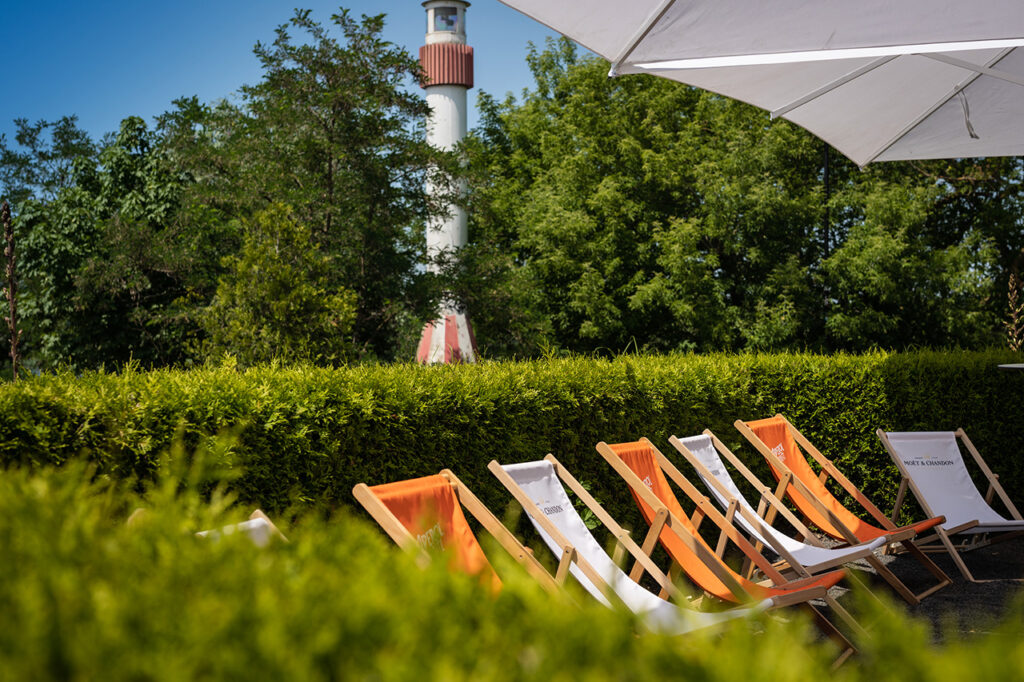 Blick auf Liegestühle vor grünen Hecken im Außenbereich des amSee, im Hintergrund der rot-weiße Leuchtturm vor der Location.