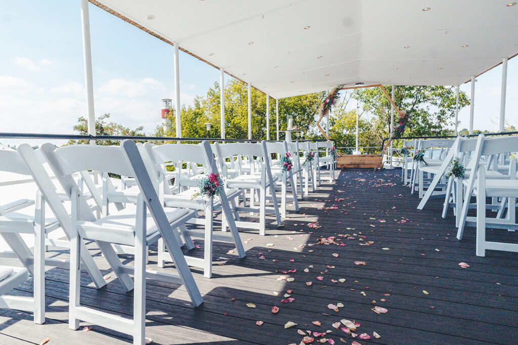 Die Oberdeck-Terrasse des amSee mit Bestuhlung für eine Freie Trauung. Weiße Vintagestühle, hölzerner Traubogen und Blumensäulen. Im Hintergrund ist der Leuchtturm zu sehen.