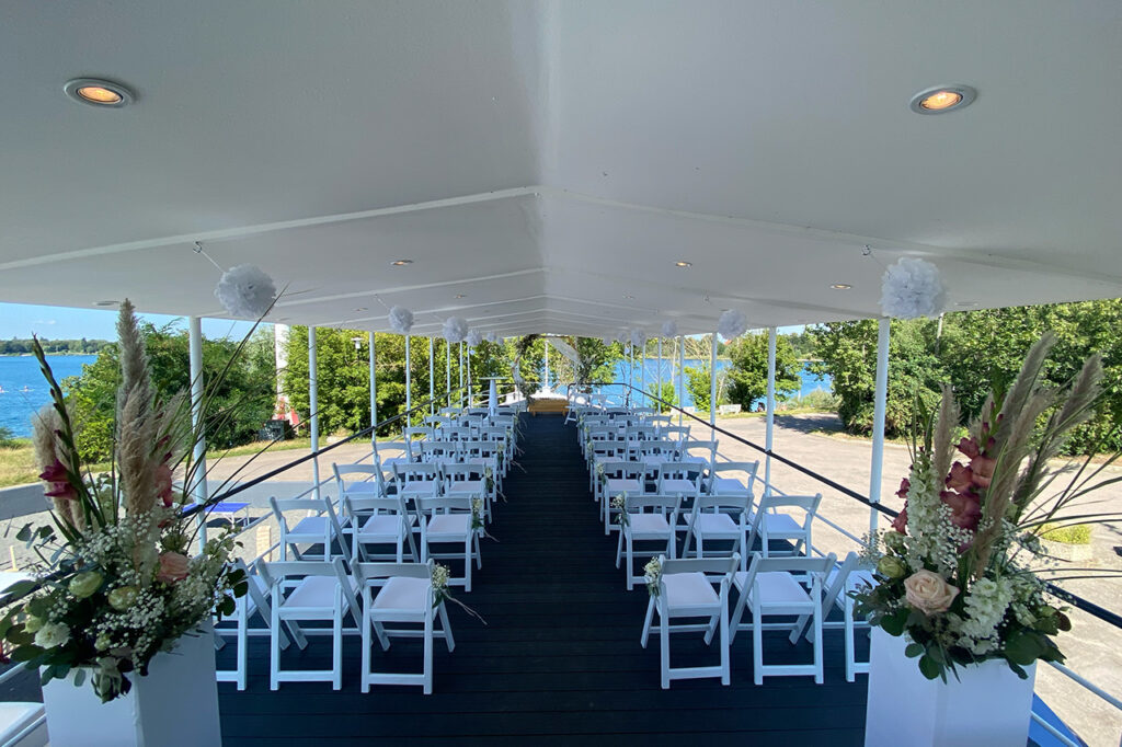 Die Oberdeck-Terrasse des amSee mit Bestuhlung für eine Freie Trauung. Weiße Vintagestühle, hölzerner Traubogen und Blumensäulen. Im Hintergrund leuchtet der See blau in der Sonne.