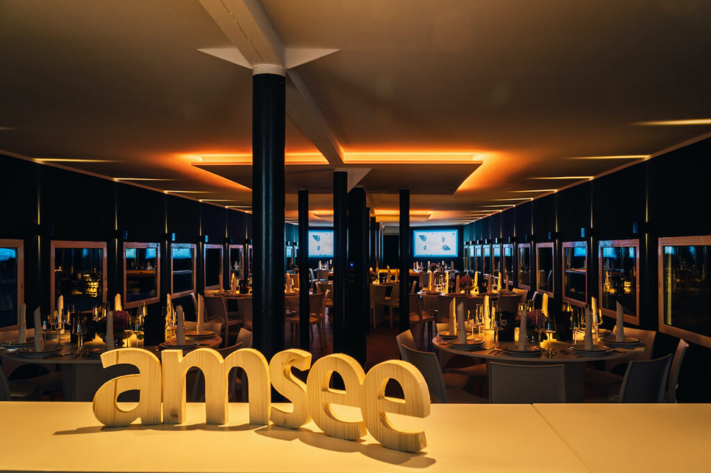 Das Oberdeck der Location amSee am Abend, die amberfarbene Beleuchtung taucht den Raum in ein schönes Licht. Im Hintergrund leuchten die zwei großen Displays an der Außenwand zur Terrasse, im Vordergrund stehen Holzbuchstaben auf der Bar: "amsee".