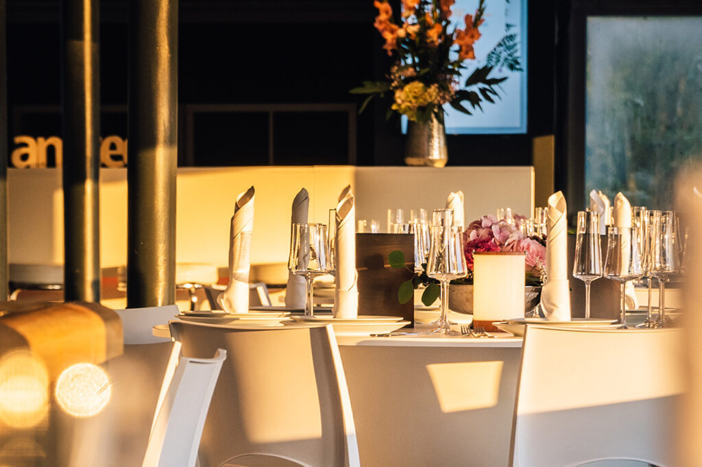 Nahaufnahme eines gedeckten Tischs im Oberdeck, im Hintergrund die Bar mit Blumenschmuck. Der Sonnenuntergang sorgt für goldenes Licht.