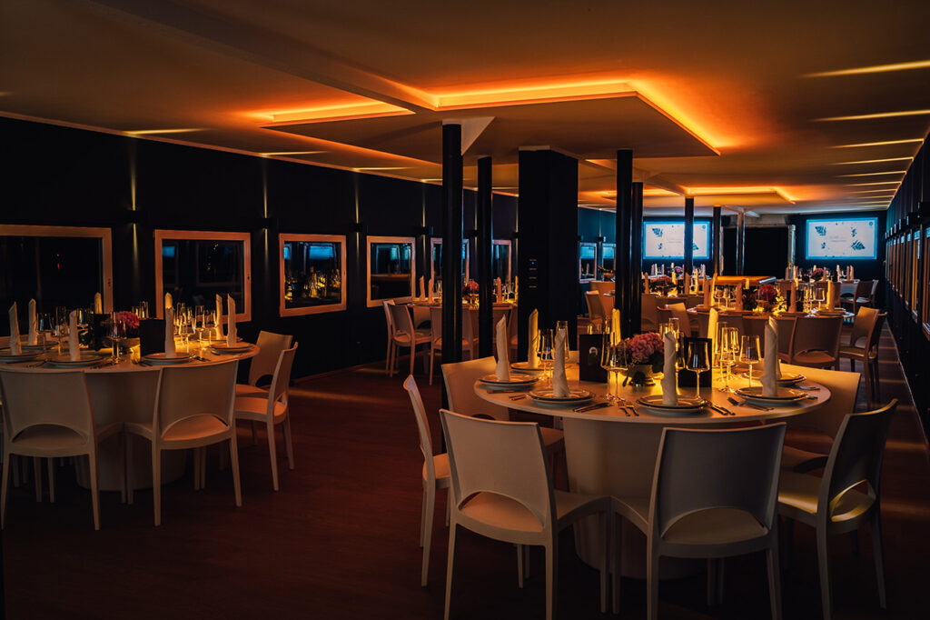Das Oberdeck der Location amSee am Abend, die amberfarbene Beleuchtung taucht den Raum in ein schönes Licht. Im Hintergrund leuchten die zwei großen Displays an der Außenwand zur Terrasse.