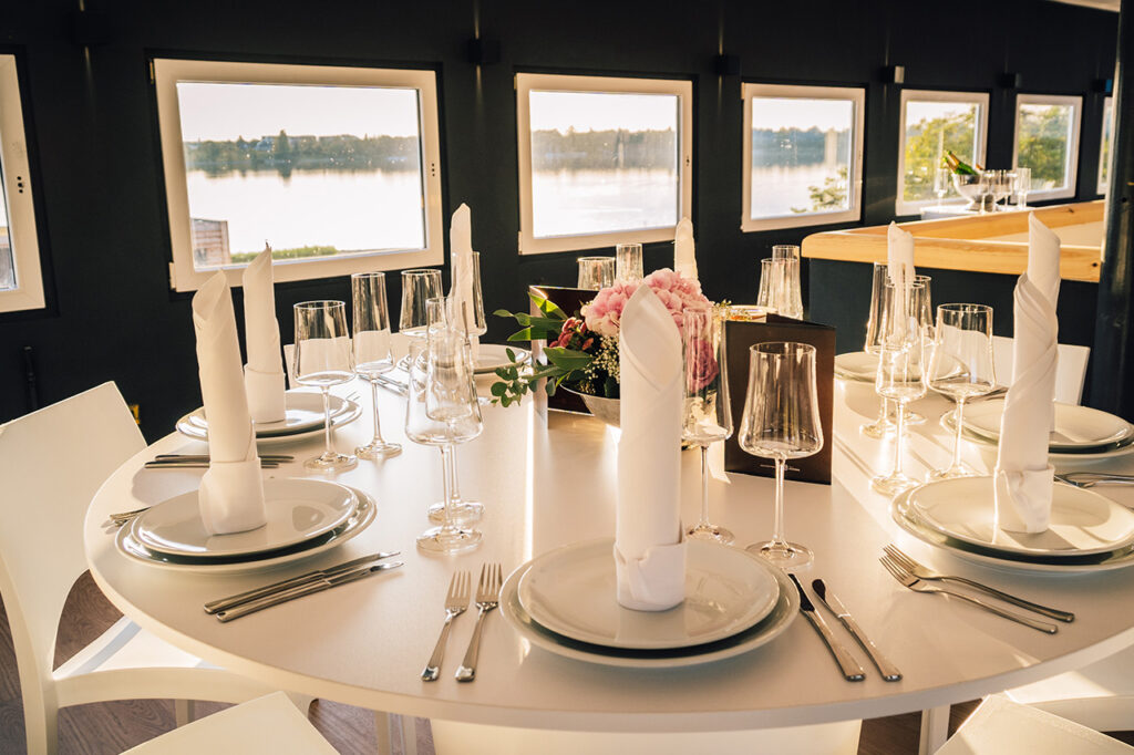 Nahaufnahme eines gedeckten Tischs im Oberdeck, im Hintergrund Bäume und der See.