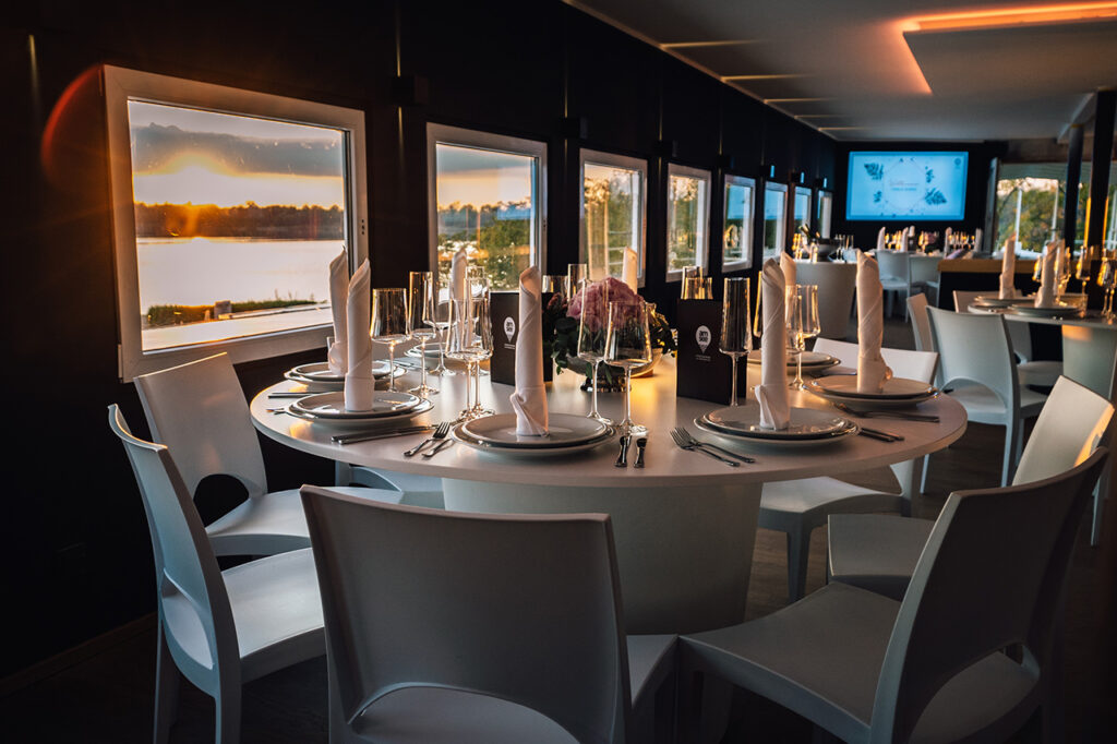 Westseite des Oberdecks mit festlichen weißen Möbeln und einem der Displays an der hinteren Wand. Durch die Fenster auf der linken Seite leuchtet der Sonnenuntergang.