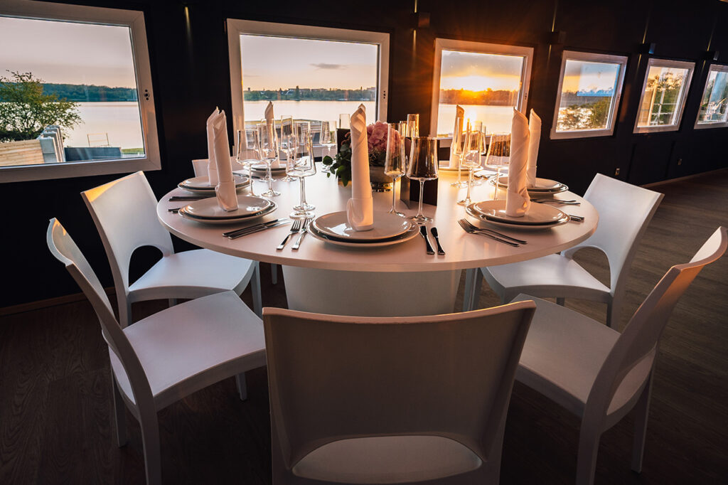 Das Oberdeck der Eventlocation amSee mit weißen modernen Möbeln. Im Hintergrund öffnet sich der Blick durch die Fenster über den von der untergehenden Sonne beschienenen See.