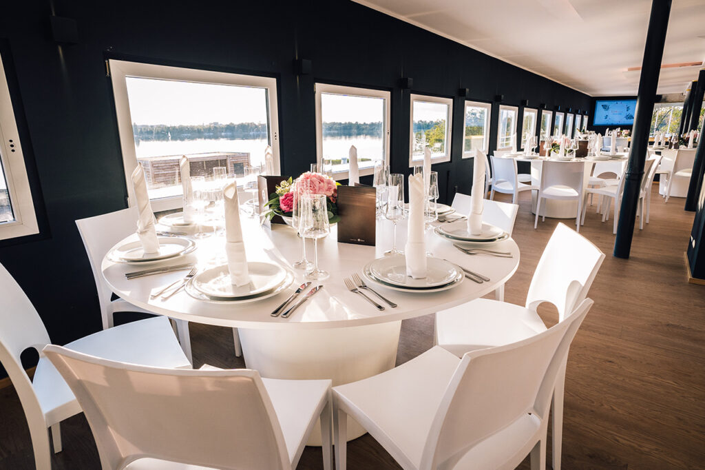 Das Oberdeck der Eventlocation amSee mit weißen modernen Bankettmöbeln. Im Hintergrund öffnet sich der Blick durch die Fenster über den von der Sonne beschienenen See.