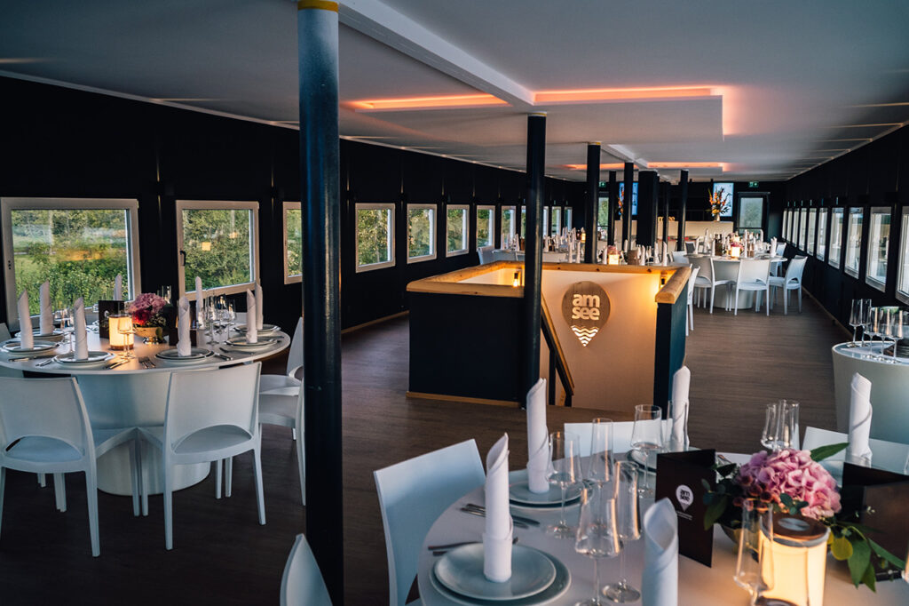 Das Oberdeck der Eventlocation amSee ist eingerichtet mit weißen modernen Bankettmöbeln. Auf den Tischen stehen Blumenschmuck, Geschirr, Gläser sowie Stoffservietten gefaltet als Kerze.