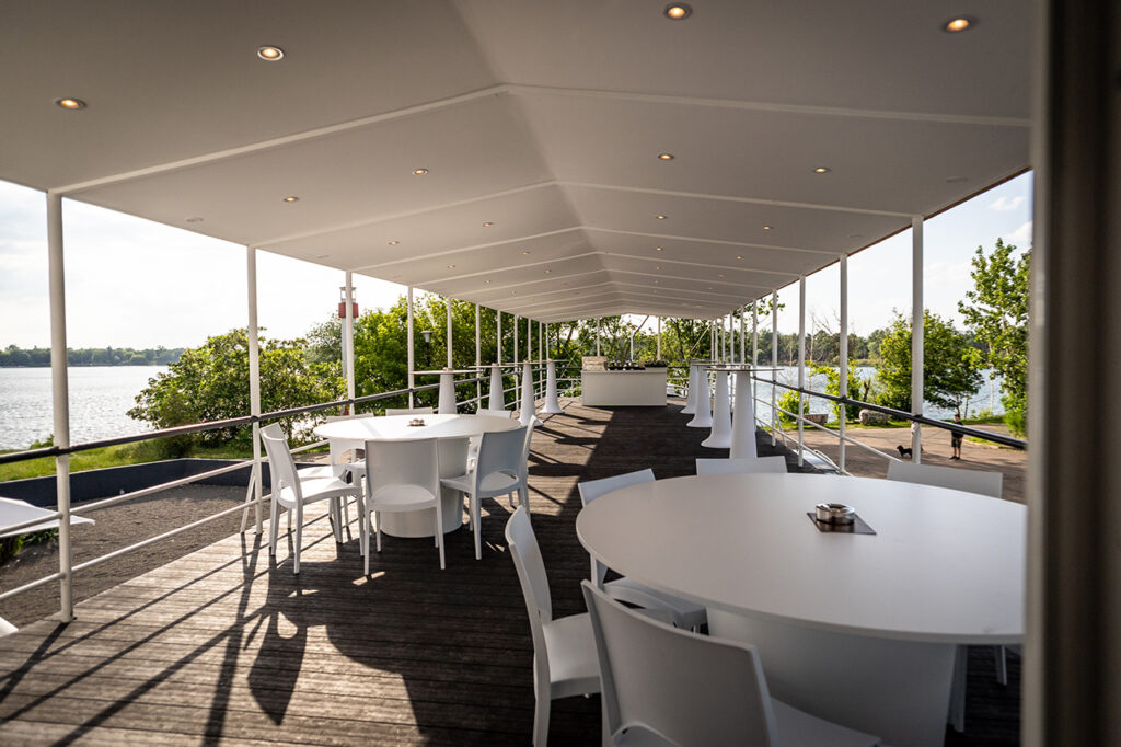 Die Oberdeck-Terrasse mit Blick auf den See und grün belaubte Bäume. Im Hintergrund befindet sich der Leuchtturm. Eingerichtet ist die Terrasse mit runden Banketttischen und -stühlen sowie Stehtischen und einer Bar in Weiß.