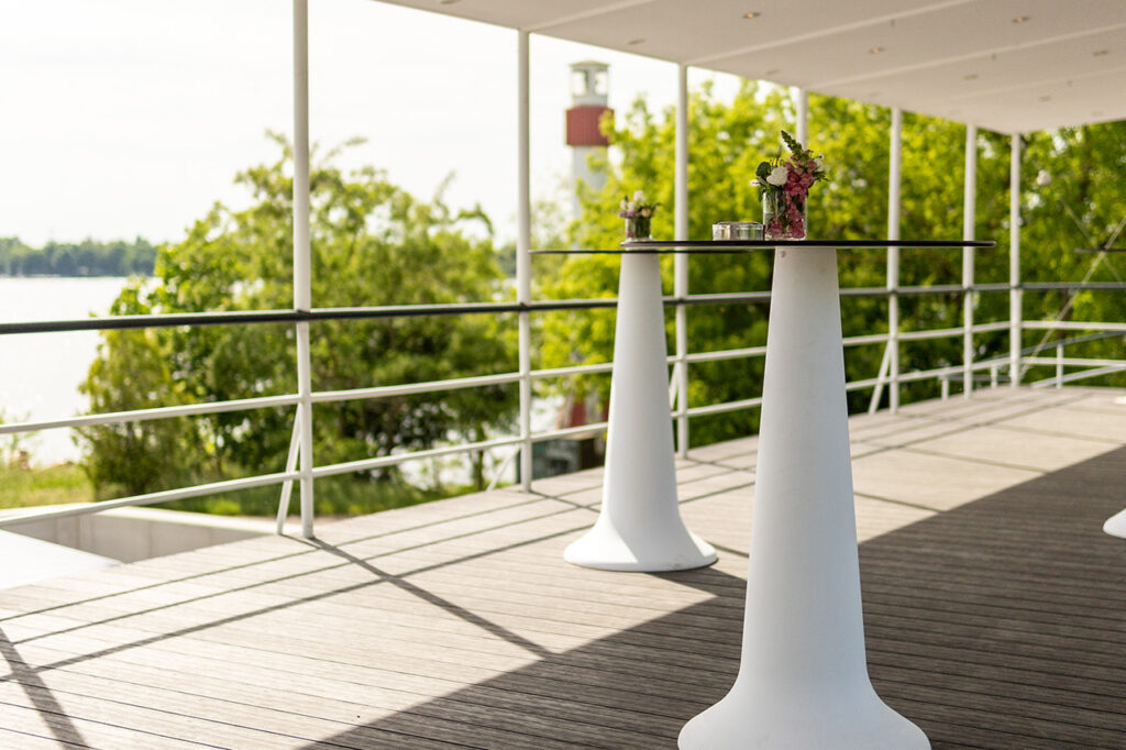 Die Oberdeck-Terrasse mit Blick auf den See und grün belaubte Bäume. Im Hintergrund befindet sich der Leuchtturm, im Vordergrund stehen zwei weiße Stehtische.