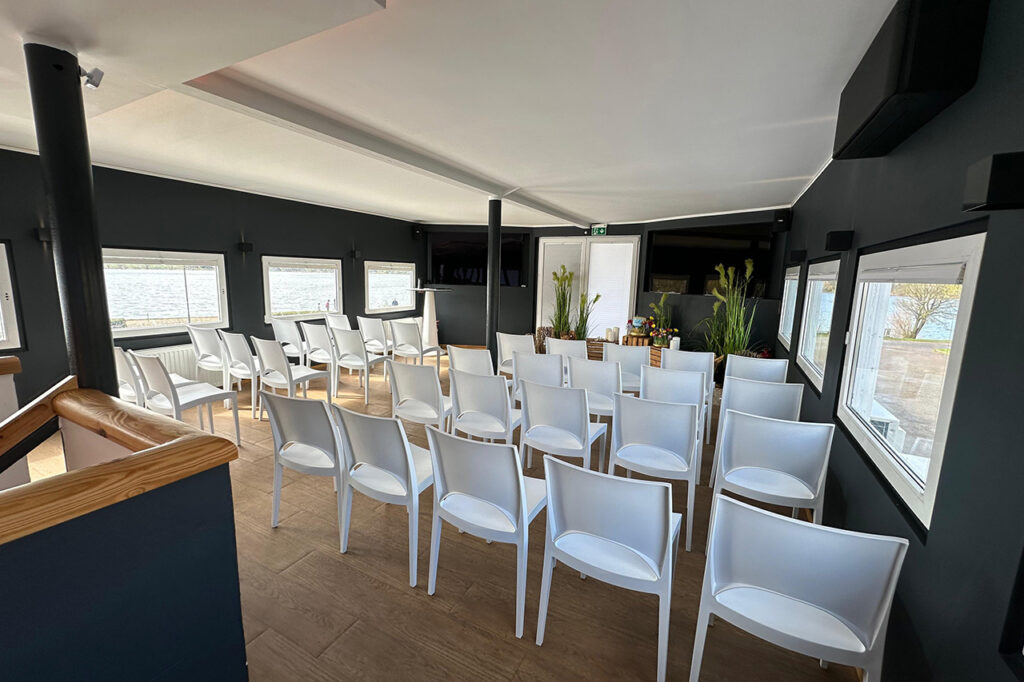 Das Oberdeck der amSee Location, eingerichtet für eine Trauerfeier: weiße Bankettstühle in Reihen sowie Holzkisten, Sand und Gräser.