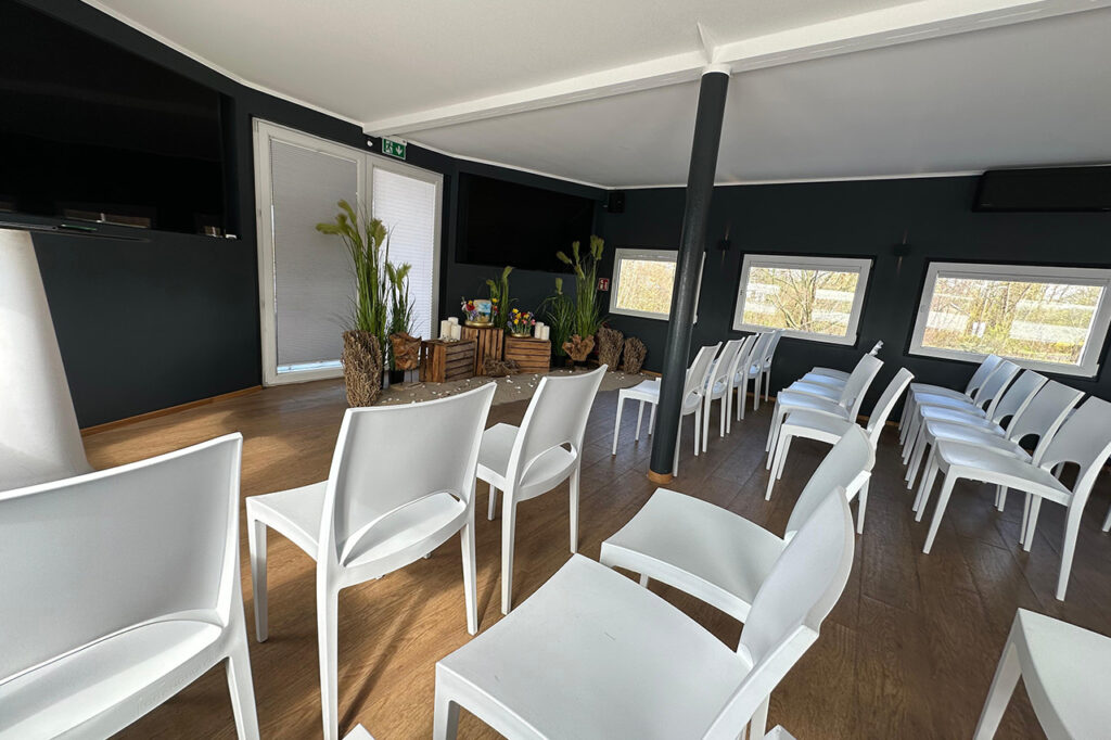 Das Oberdeck der amSee Location, eingerichtet für eine Trauerfeier: weiße Bankettstühle in Reihen sowie Holzkisten, Sand und Gräser.