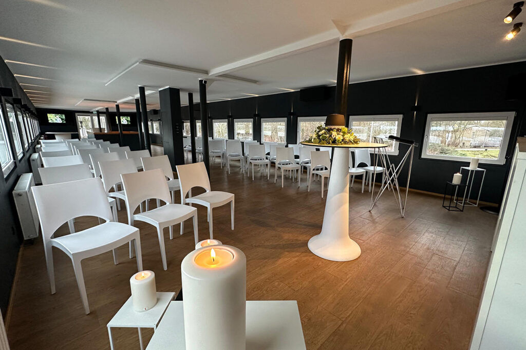 Das Oberdeck der amSee Location, eingerichtet für eine Trauerfeier: weiße Bankettstühle in Reihen sowie ein weißer Stehtisch mit Urne und Blumenschmuck.