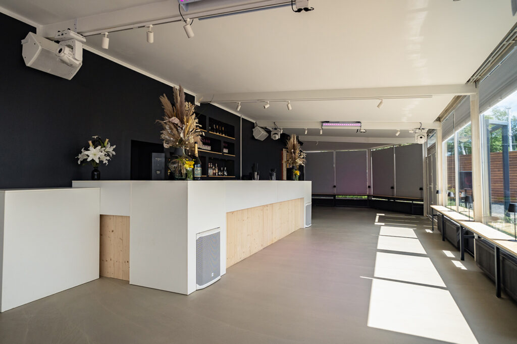 Der Wintergarten mit der großen Hauptbar, vom Eingang aus fotografiert. Für Akzente sorgen die schwarze Wand hinter der Bar sowie der Tresen in Weiß mit Holzelementen.