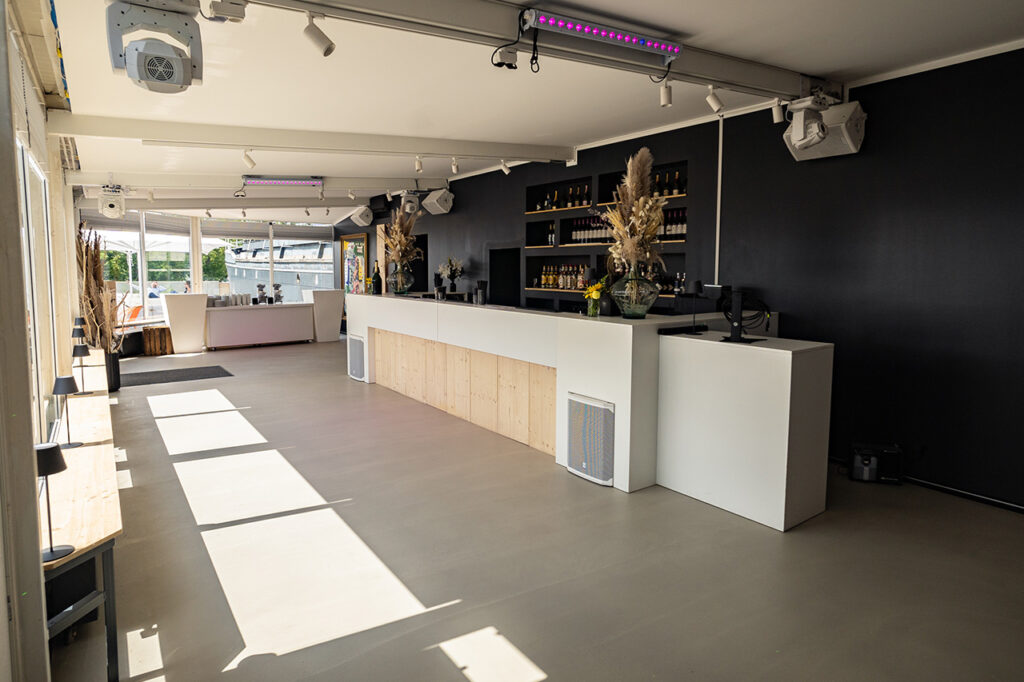 Der Wintergarten mit der großen Hauptbar. Für Akzente sorgen die schwarze Wand hinter der Bar sowie der Tresen in Weiß mit Holzelementen.