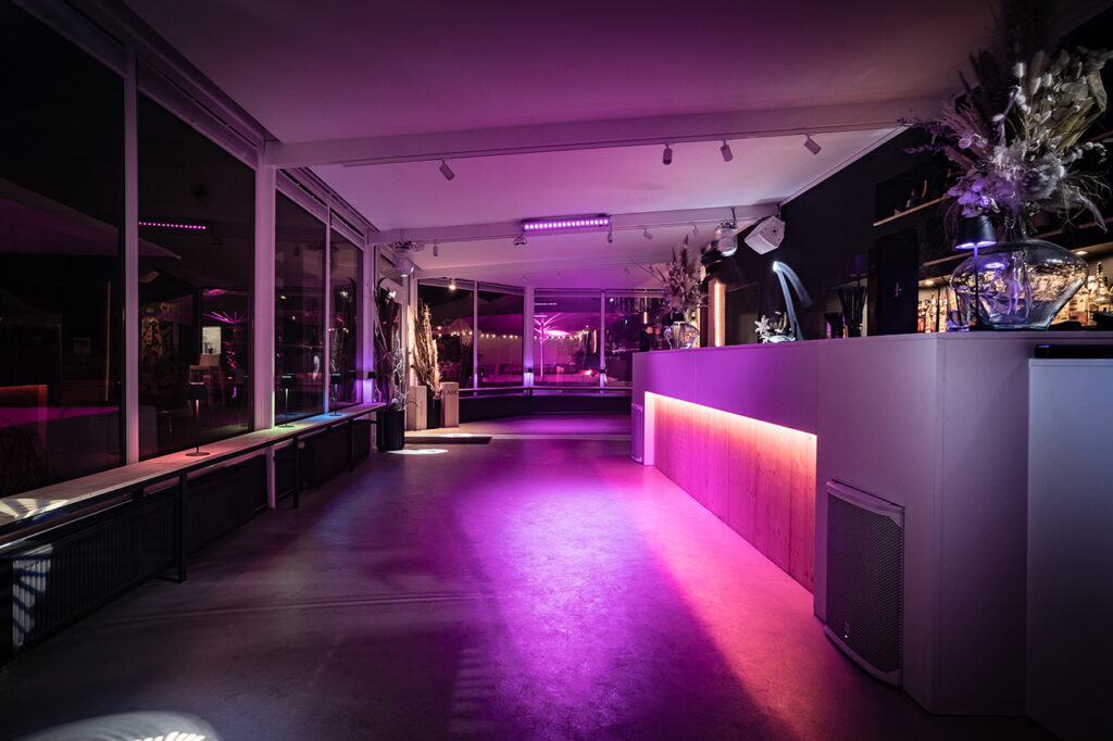 Der Wintergarten der Eventlocation amSee am Abend. Draußen ist es dunkel, lilafarbenes und bernsteinfarbenes Licht sorgt für Partystimmung.
