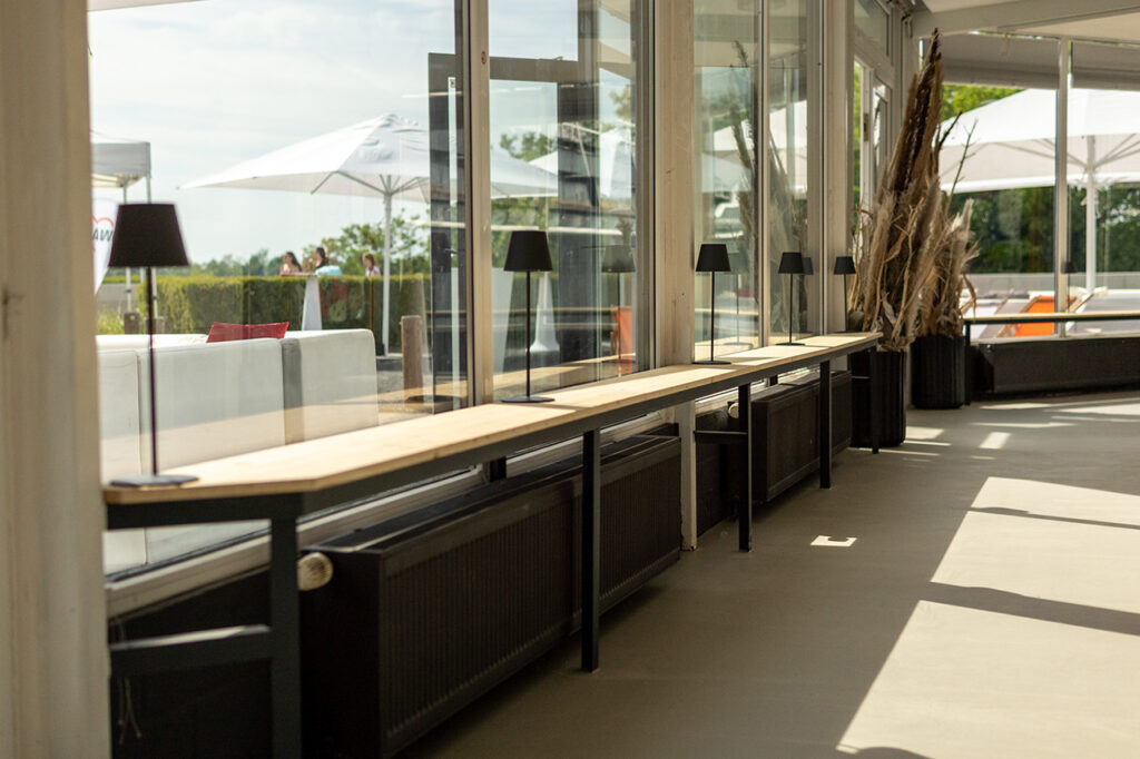 Blick aus dem Wintergarten in Richtung Außenbereich und See. Im Vordergrund befinden sich niedrige Sitzbänke aus schwarzem Stahl und Holz mit schwarzen kleinen Ambientelampen.