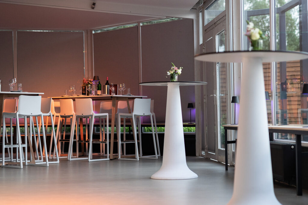 Teilaufnahme des Wintergartens der Location mit zwei modernen weißen Stehtischen und im Hintergrund Sitzgruppen aus Hochtischen und Barhockern.