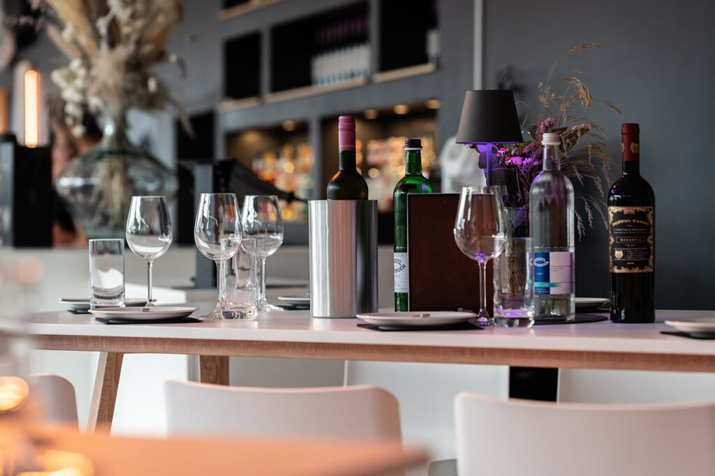Nahaufnahme eines Tischs vor der Bar im Wintergarten. Darauf stehen mehrereTeller, Weingläser sowie verschiedene Flaschen Wein und Wasser.