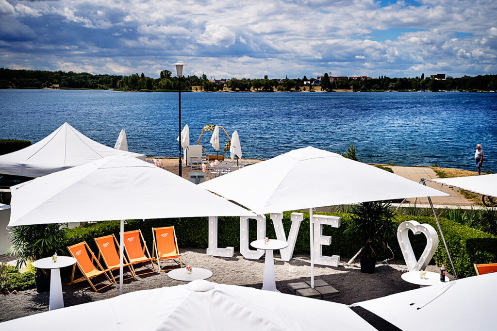 Das Bild zeigt ein mögliches Setting für eine Hochzeit im amSee: Auf der Sandterrasse ist stehen Stühle und ein Traubogen für eine freie Trauung, der Außenbereich vor der Location ist mit Liegestühlen, Leuchtbuchstaben und Sonnenschirmen möbliert.