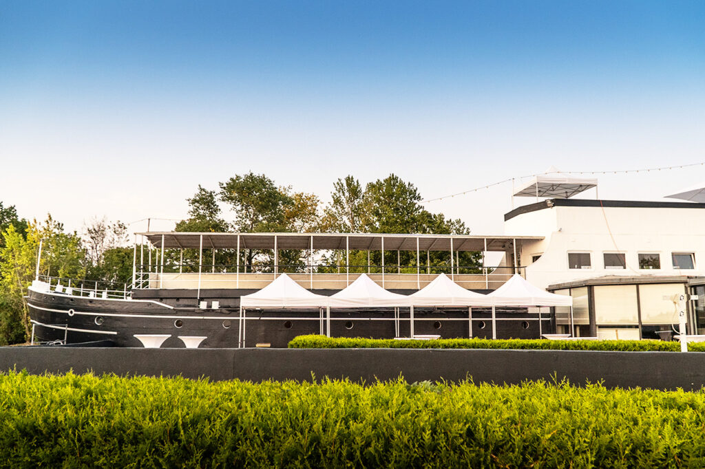 Die amSee Location aufgenommen vom Seeufer aus. Das Schiff ist in Schwarz und Weiß gestrichen.