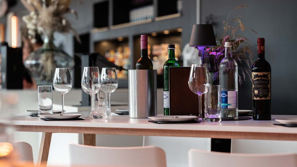 Nahaufnahme eines Tischs vor der Bar im Wintergarten. Darauf stehen mehrereTeller, Weingläser sowie verschiedene Flaschen Wein und Wasser.