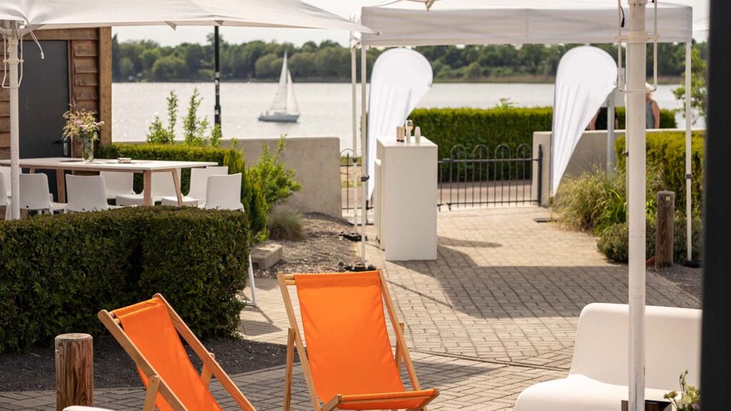 Liegestühle und weitere Sitzmöglichkeiten im Außenbereich der amSee Location, im Hintergrund ein Segelschiff auf dem See.